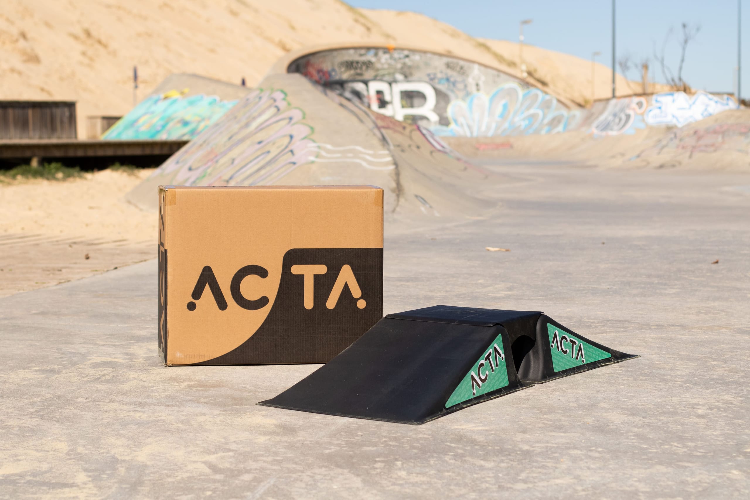 Carton ACTA et rampe noire, posés dans un skatepark extérieur