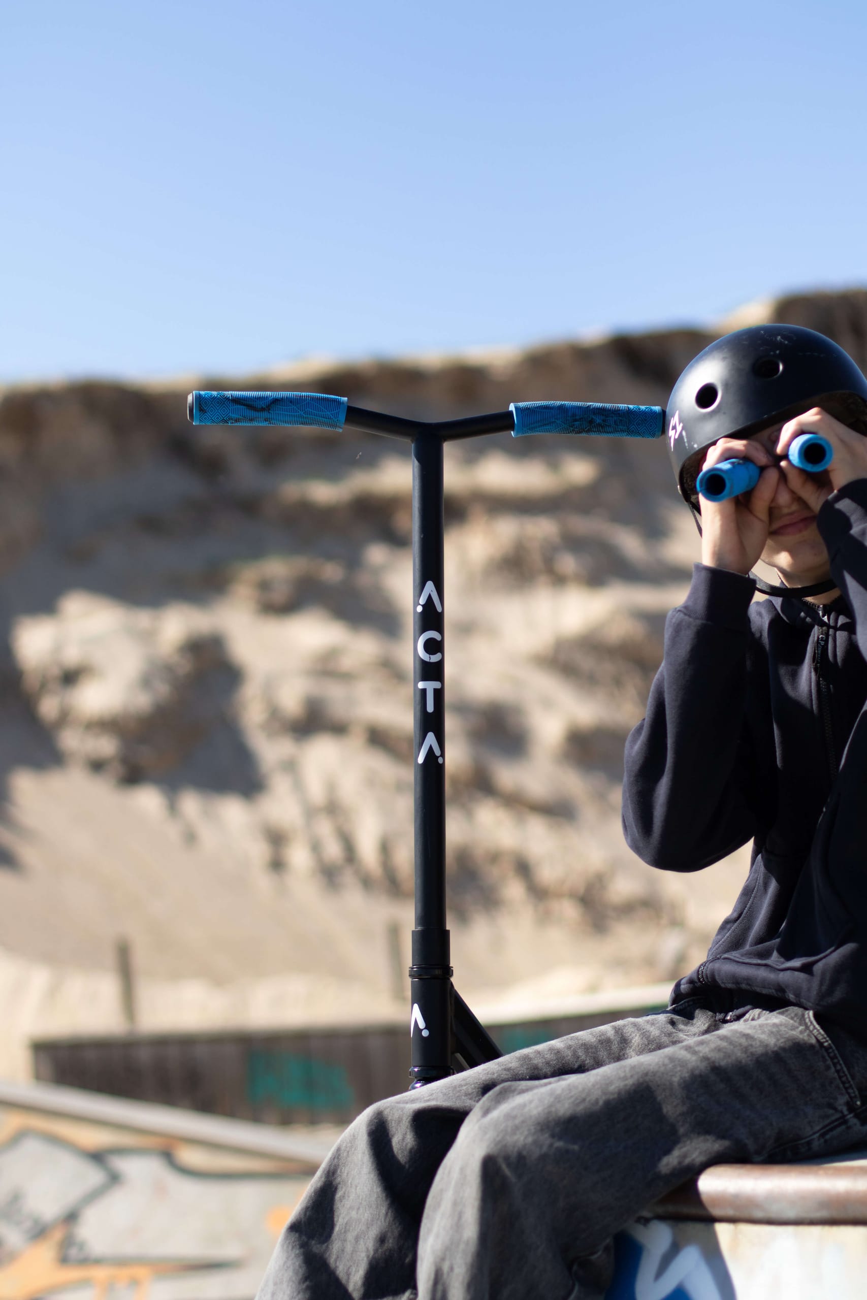 Guidon de trottinette freestyle ACTA avec poignées bleues, rider en arrière-plan