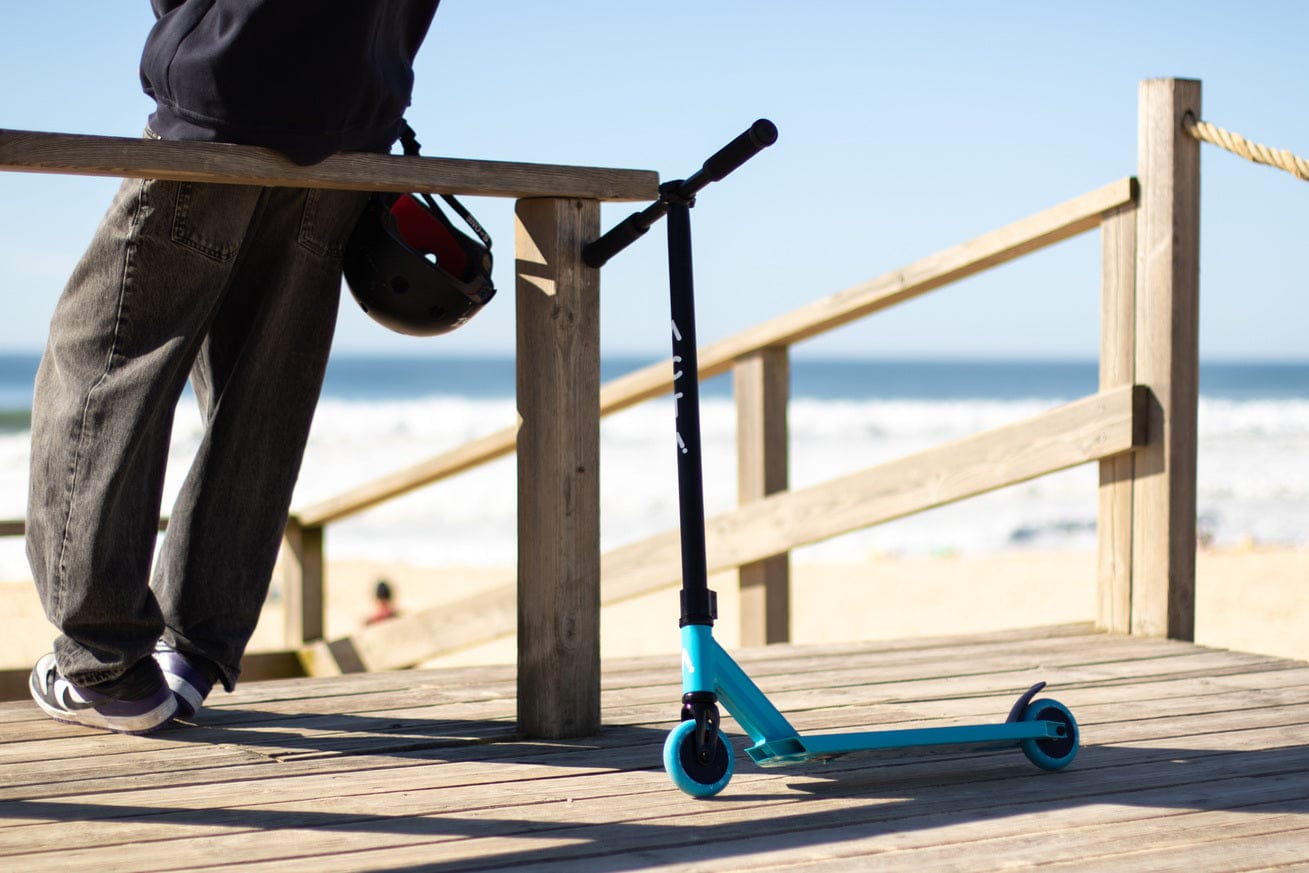 Trottinette freestyle ACTA Teal posée sur une terrasse en bois, plage en arrière-plan