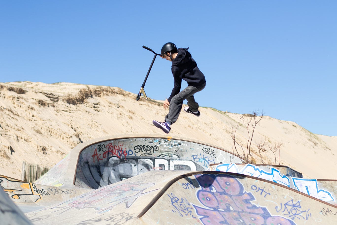 Rider en trottinette freestyle sautant au-dessus d'un module de skatepark tagué, dunes en arrière-plan