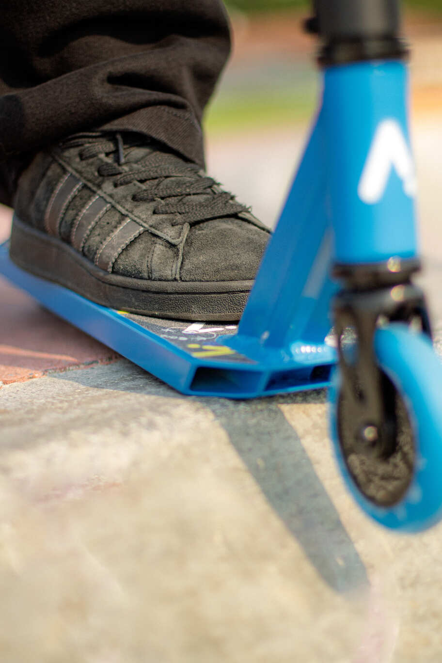 Chaussure posée sur le deck d'une trottinette freestyle ACTA bleue, gros plan sur la roue